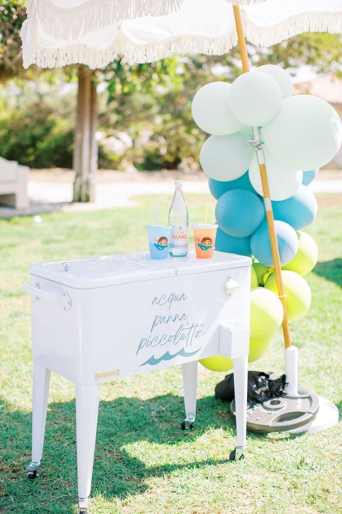 Weiße Kühlbox mit Flaschen, bunten Bechern und Luftballons, sonniger Tag im Park.